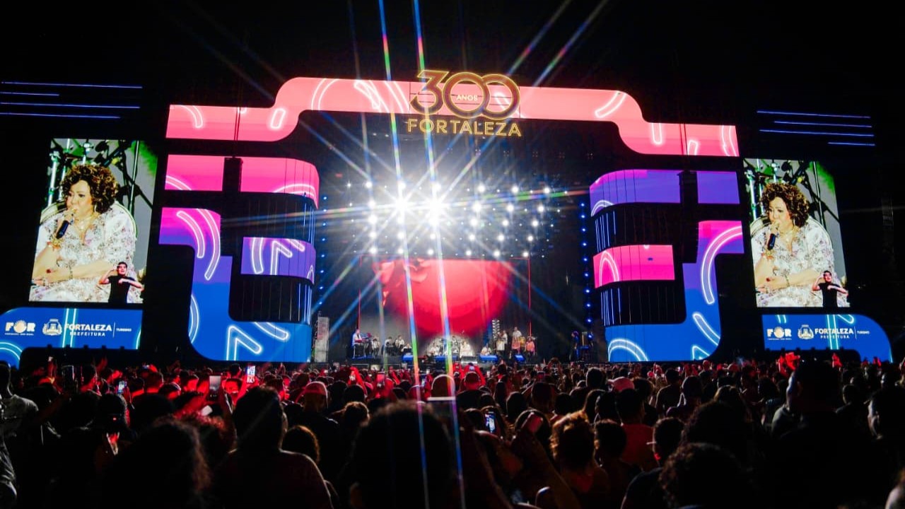 A foto mostra o palco dos shows do tricentenário de Fortaleza no Aterrinho da Praia de Iracema. Em primeiro plano, no contraluz, há uma multidão.